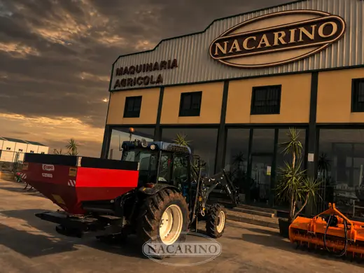 Tractor azul con abonadora roja delante de la nave de Nacarino en Aceuchal