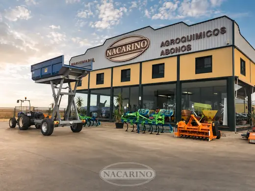 Remolque elevador de tijera azul con la caja elevada, junto a un tractor delante de la nave de Nacarino en Aceuchal