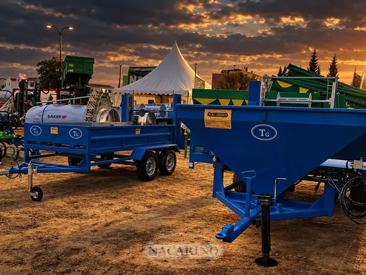 Remolque plataforma azul cargado con un equipo de tratamientos y tolva azul en una feria agrícola, al atardecer
